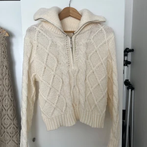 Vit stickad tröja med dragkedja - Säljer en mysig vit stickad tröja med dragkedja och hög krage. Tröjan har ett snyggt flätmönster och ribbade muddar vid ärmslut och nederkant. Perfekt för kyliga dagar!