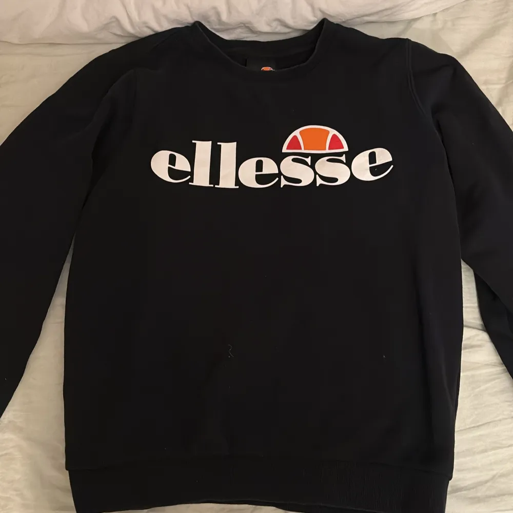 Säljer en svart tröja från Ellesse med deras ikoniska logga i vitt och orange på bröstet. Tröjan har långa ärmar och en rund halsringning. Perfekt för en avslappnad stil.. Neuletakit & Villapaidat.