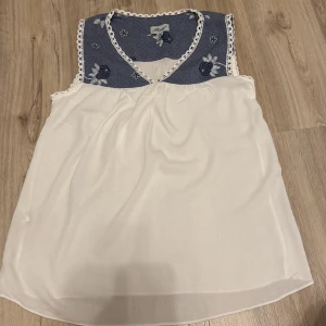 Vit och blå ärmlös blus från Forel - Säljer en söt ärmlös blus från Forel i vitt med blå detaljer. Blusen har en v-ringad design med spetskant och ett blommigt mönster på den blå delen. Perfekt för en somrig look! Made in europe!
