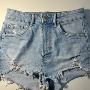 Ljusa jeansshorts från Zara - Säljer ett par ljusa jeansshorts från Zara med fransiga kanter och slitna detaljer. Shortsen har en klassisk femficksdesign och knappgylf. Perfekta för en avslappnad sommarlook! 🌞