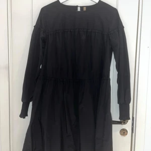 Svart klänning med volangdetaljer - Elegant svart klänning med volangdetaljer och långa ärmar. Perfekt för en stilren look. Klänningen har en lös passform och är tillverkad i ett mjukt material.