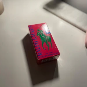 Ralph Lauren Eau de Toilette för kvinnor - Säljer en elegant Ralph Lauren Eau de Toilette för kvinnor. Förpackningen är i en livfull rosa färg med en grön häst och ryttare samt blå text. Flaskan innehåller 50 ml av doften. Helt ny 