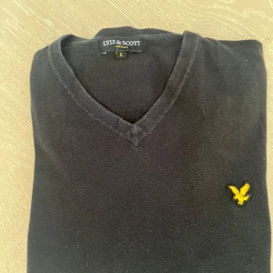Svart tröja från Lyle & Scott - Säljer en stilren svart tröja från Lyle & Scott med v-ringning och det klassiska gula logotypen på bröstet. Perfekt för en avslappnad men ändå snygg look. Strl. L. Har ett pyttelitet hål under ena armen. Rök och djurfritt hem.