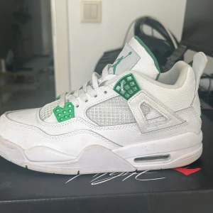 Jordans 4  - Snygga vita Nike Air Jordans med gröna detaljer. Skorna har snörning och en ikonisk Jumpman-logga i grönt på hälen och tungan. Perfekta för dig som vill ha en stilren och sportig look.