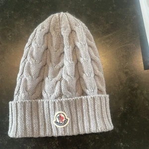Grå stickad mössa från Moncler - Snygg grå stickad mössa från Moncler med kabelstickat mönster och Moncler-logga framtill ger en exklusiv touch. Använd väldigt få gånger.