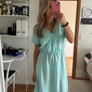 Ljusblå omlottklänning - Säljer en söt ljusblå omlottklänning med korta ärmar. Perfekt för varma dagar med sin luftiga design och elastiska midja som ger en bekväm passform. Klänningen har en v-ringad hals och är gjord i ett mjukt material.