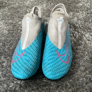 Nike fotbollsskor i blått och grått - Snygga Nike fotbollsskor i en fräsch kombination av blått och grått med rosa detaljer. Skorna har en modern design utan snörning för en strömlinjeformad look och optimal passform på planen.