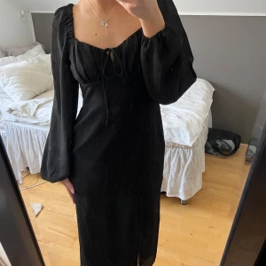 Svart långklänning med puffärmar - Elegant svart långklänning med puffiga långa ärmar och en söt knytning framtill. Klänningen har en smickrande slits och en åtsittande passform som framhäver figuren. 