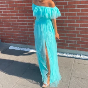 Turkos långklänning - Säljer en vacker turkos långklänning med offshoulder-design och volangdetaljer. Klänningen har en elegant slits framtill som ger en fin rörelse. Perfekt för sommarens festligheter!