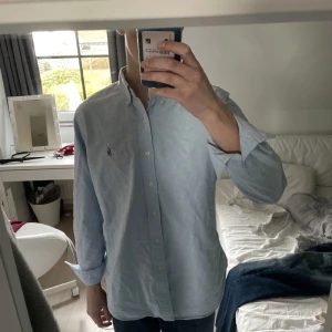 Ljusblå skjorta från Ralph Lauren - Säljer en klassisk ljusblå skjorta från Ralph Lauren med broderad logotyp på bröstet. Skjortan har långa ärmar och knappar framtill samt på kragen. Perfekt för en stilren look. Skjortan är använd ett fåtal gånger och säljer då den är för stor för mig!