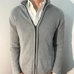 Grå stickad kofta med dragkedja - Säljer en stilren grå stickad kofta med dragkedja från Cedar Wood State. Koftan har långa ärmar och ribbade detaljer vid ärmslut och nederkant. Perfekt för kyligare dagar. Bra skick, inga defekter. Modellen är 182cm/67kg