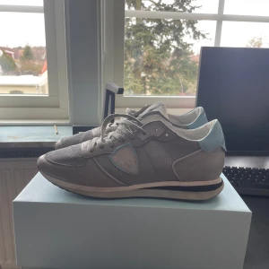 Grå sneakers från Philippe Model - Snygga grå sneakers från Philippe Model med ljusblå detaljer och snörning. Skorna har en rund tå och en bekväm sula, perfekt för vardagsbruk. Tillverkade i en kombination av skinn och syntetmaterial.