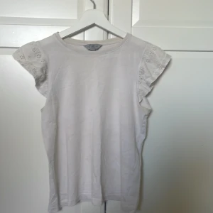 Vit topp från TIKI - Säljer en söt vit topp från TIKI med korta ärmar och spetsdetaljer. Perfekt för varma dagar och enkel att matcha med olika outfits.