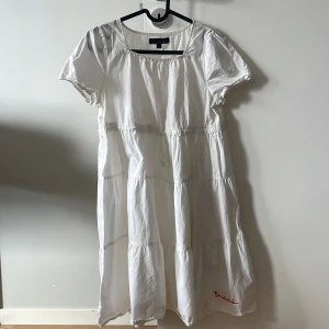 Vit klänning med broderad text - Charmig vit klänning med korta ärmar och broderad text i rött på baksidan. Klänningen har en luftig passform och är perfekt för varma dagar. Den har en söt detalj med ett hjärta bredvid texten. Fin för en midsommar dag🥰 (passar även barn som har storlek 160)