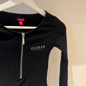 Svart långärmad topp från Guess - Snygg svart långärmad body från Guess med dragkedja framtill och logga på bröstet. Perfekt för en stilren look. Tillverkad i ett mjukt material som ger en bekväm passform.  Tröjan är köpt på Sicilien i Italien. Ny pris: 600