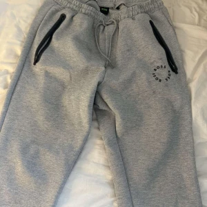 Grå mjukisbyxor från Boss - Snygga grå mjukisbyxor från Boss med elastisk midja och dragsko. Byxorna har svarta dragkedjor på fickorna och en diskret logga på benet. Perfekta för en avslappnad stil.