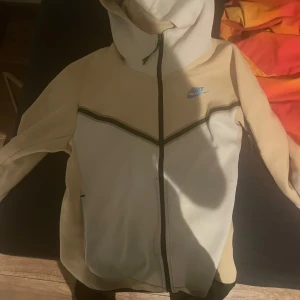 Beige och vit nike tech från Nike - Säljer en snygg beige och vit hoodie från Nike med dragkedja och huva. Den har en blå Nike-logga på bröstet och svarta detaljer längs dragkedjan och ärmarna. Perfekt för en sportig look!