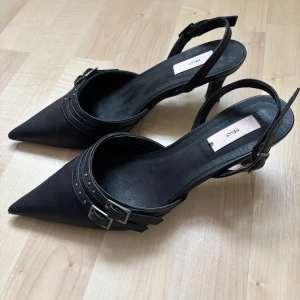 Pointy buckle heel - Snygga svarta pumps från Nelly med spetsig tå och klack. Skorna har dubbla spännen och dekorativa nitar för en stilren look. Perfekta för en elegant touch till din outfit. Klackhöjd: 4cm. Storlek 37 men passar mig som brukar ha 38. Nypris: 399kr
