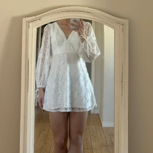 Vit klänning  - Säljer en söt vit spetsklänning med v-ringning och långa ärmar. Klänningen har ett fint mönster och knytband vid ärmsluten. Perfekt för sommarens alla tillfällen! 💫