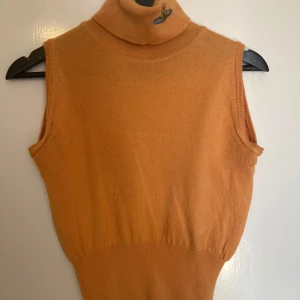 Orange ärmlös polotröja från Vivienne Westwood - Säljer en snygg orange ärmlös polotröja från Vivienne Westwood Red Label. Tröjan har en hög krage med en broderad detalj och är perfekt för lager-på-lager-stil. Den är tillverkad i ett mjukt material som ger en skön passform.Eftersom den är vintage finns det urnoppade ställen men inget som verkligen sticker ut 