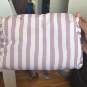 Randig necessär i lila och vit - Säljer en söt necessär med lila och vita ränder. Den har en praktisk dragkedja och är perfekt för att förvara dina småsaker. Insidan är lila och rymlig nog för dina behov. Har några fläckar men det kommer tvättas ut innan jag skickar ut 