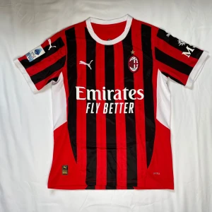 AC Milan fotbollströja - Säljer en AC Milan fotbollströja från Puma med klassiska röda och svarta ränder. Tröjan har korta ärmar och vit krage. Den har sponsorloggan 'Emirates Fly Better' på framsidan och klubbmärket med en stjärna ovanför. Perfekt för fans av laget!