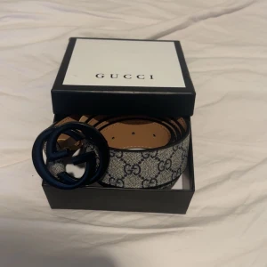 Gucci bälte med GG-mönster - Snyggt Gucci bälte med det klassiska GG-mönstret i beige och svart. Bältet har en rund svart spänne med GG-logotypen. Perfekt för att ge en lyxig touch till din outfit.