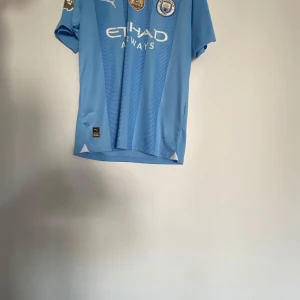 Blå fotbollströja från Puma - Säljer en blå fotbollströja från Puma med korta ärmar. Tröjan har ett tryck på framsidan med sponsorlogga och klubbmärke. På baksidan finns namnet 'De Bruyne' och nummer 17. Perfekt för fotbollsfans! Storlek M