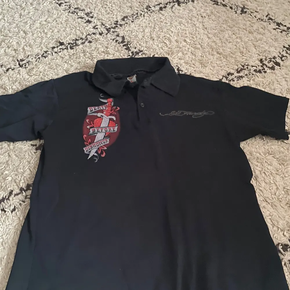 Säljer en svart kortärmad t-shirt från Ed Hardy med ett stort tryck på ryggen som säger 'Death Before Dishonor'. T-shirten har en klassisk krage och är gjord i bomull. Perfekt för dig som gillar en edgy stil!. T-paidat.