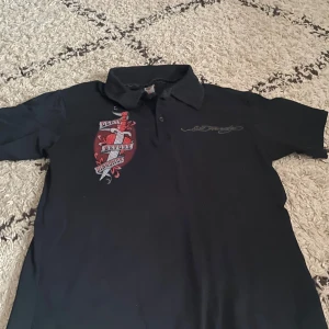  Ed Hardy - Säljer en svart kortärmad t-shirt från Ed Hardy med ett stort tryck på ryggen som säger 'Death Before Dishonor'. T-shirten har en klassisk krage och är gjord i bomull. Perfekt för dig som gillar en edgy stil!