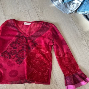 Röd mönstrad blus från Vila - Säljer en snygg röd blus från Vila med ett detaljerat blommönster i mörkare nyanser. Blusen har en v-ringad hals och långa ärmar med volangdetaljer vid ärmsluten. Perfekt för att ge en färgklick till din outfit! Är lite genomskinlig🩷🩷