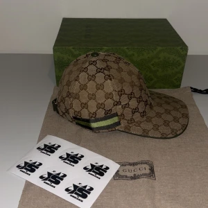 Beige keps från Gucci - Snygg beige keps från Gucci med det klassiska GG-mönstret. Kepsen har gröna och svarta ränder på sidan och justerbar rem baktill. Perfekt för att ge din outfit en lyxig touch. Finns i S & M