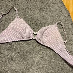 Lila och vit rutig bikini set - Söt bikini i lila och vitt med rutigt mönster. Toppen har trekantsform och underdelen har dekorativa hjärtdetaljer på sidorna. Överdelen strl S och underdel strl XS