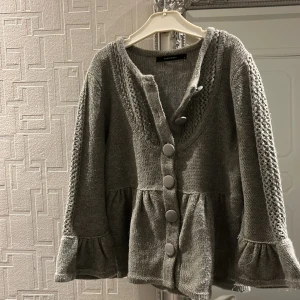 Grå stickad kofta med volangdetaljer - Säljer en mysig grå stickad kofta med stora knappar och volangdetaljer vid ärmslut. Perfekt för kyliga dagar och sommarkvällar. Passar bra till både jeans och kjol.