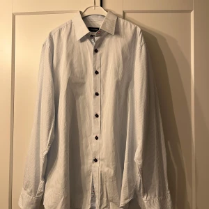 Vit och blå randig skjorta från Dressmann - Säljer en stilren vit och blå randig skjorta från Dressmann Premium Collection. Skjortan är tillverkad i tvåskikts egyptisk bomull och har långa ärmar med knappar. Perfekt för en elegant look. KNAPPAST ANVÄND!!