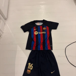 FC Barcelona matchtröja med shorts - Säljer en FC Barcelona matchtröja med tillhörande shorts. Tröjan har klassiska röda och blå vertikala ränder, klubbmärke, Nike-logga och Spotify som sponsor framtill. Nummer 16 i guld på shortsen och nummer 10 med UNHCR-logga på ryggen. Materialet är lätt och andas.