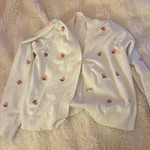 Vit kofta med broderade blommor - Söt vit kofta med broderade små blommor i rosa och gult över hela tröjan. Koftan har ribbade muddar, knappar framtill och långärmad passform. Perfekt för dig som gillar gulliga detaljer och vill ha något unikt i garderoben.