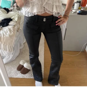 Svarta bootcut byxor med fickdetaljer - Snygga svarta byxor med bootcut-modell och dekorativa fickor framtill. Byxorna har två knappar i midjan och en lätt glansig finish som ger en cool look. Perfekta att styla med crop tops eller stickade tröjor.
