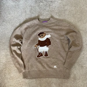 Purple Label Ralph Lauren Polo Bear i Kaschmir - Säljer en beige stickad tröja från Ralph Lauren med en stor broderad björn på framsidan. Tröjan har rund hals, ribbade muddar och RL-broderi vid nederkanten. Tillverkad i mjuk kashmir, perfekt för dig som vill ha något extra nice och unikt i garderoben. Hittades på myrorna för några veckor sedan men krympte i tvätten och är nu som ett S istället för L. 