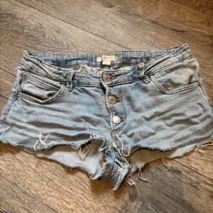 Ljusblå jeansshorts från Gina Tricot - Snygga ljusblå jeansshorts från Gina Tricot med fransig och rå kant, klassisk femficksdesign och knappgylf med tre silverfärgade knappar. Modellen är lågmidjad och har en loose passform, perfekt för chill dagar under sommaren.