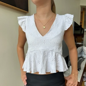 Vit blus med volang och broderi - Superfin vit blus med v-ringning, volang nertill och broderade hålmönster. Ärmlös med volangiga axlar och lätt strukturerat tyg som känns luftigt. 