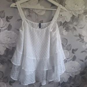 Vit volangtop från H&M Divided - Superfin vit linnetopp från H&M Divided med flera lager volanger och små broderade prickar. Toppen är ärmlös med breda axelband och har en luftig, flowy känsla – perfekt för varma dagar eller när du vill ha en söt och romantisk look.