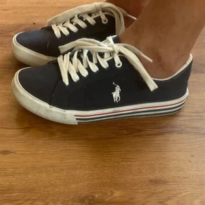 Mörkblå sneakers från Polo Ralph Lauren st38 - Snygga mörkblå sneakers från Polo Ralph Lauren med vit logga på sidan. Skorna har vita snören, vit sula med röd och blå rand samt klassisk låg profil. I väldigt bra skick. St 38