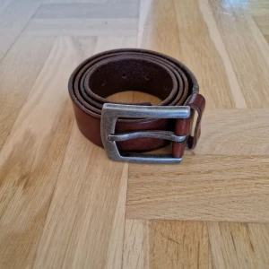 Brunt skärp från Jean Paul i skinn - Snyggt brunt bälte från Jean Paul med präglad logga och robust metallspänne. Tillverkat i äkta skinn och har en klassisk rak form. Perfekt för dig som vill ha en stilren accessoar med lite vintagekänsla. Längden är cirka 117 cm från ände till ände och storlek 90.
