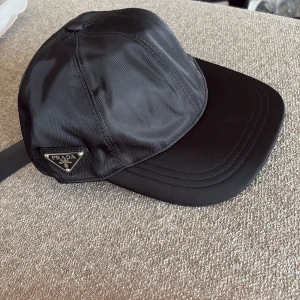 Svart keps från Prada i nylon - Snygg svart keps från Prada med klassisk böjd skärm och ikonisk metallogo på sidan. Tillverkad i glansigt nylonmaterial som ger en lyxig känsla. Perfekt accessoar för att lyfta din outfit och visa din stil.