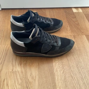 Svarta sneakers från Philippe Model - Snygga svarta sneakers från Philippe Model med detaljer i mocka och skinn samt silvriga partier vid hälen. Skorna har snörning och en platt sula. Perfekta för dig som gillar en stilren look. En skada (se på bild 3) på sidan av vänstra skon, men som enkelt går att fixa.
