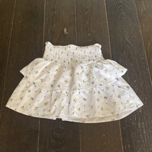 Vit blommig volangkjol Gina Tricot - vit blommig kjol från Gina Tricot med små blommor i blått och gult. Kjolen har smock i midjan och dubbla volanger. Både söt och bekväm!!