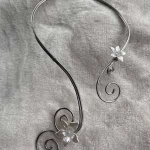 Silvrigt halsband med blommor och spiraler - Unikt halsband i silverfärgad metall med två blommor och snygga spiralformade detaljer. Blommorna har en pärlemorskimrande finish som ger en extra touch. Halsbandet ligger snyggt runt halsen och är perfekt för dig som gillar statement-smycken med lite magisk vibe.