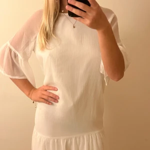 Vit kortklänning med volangärm - Säljer en söt vit kortklänning med transparenta volangärmar och volangkant nedtill. Klänningen har en loose passform och är tillverkad i ett lätt, luftigt material som ger en fräsch och somrig vibe. 