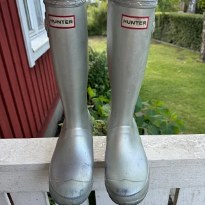 Silvriga Hunter stövlar - Säljer ett par silvriga Hunter stövlar med klassisk hög modell och justerbart spänne på sidan. Stövlarna är tillverkade i syntetmaterial och har rund tå samt platt sula. Perfekta för regniga dagar och festivaler.